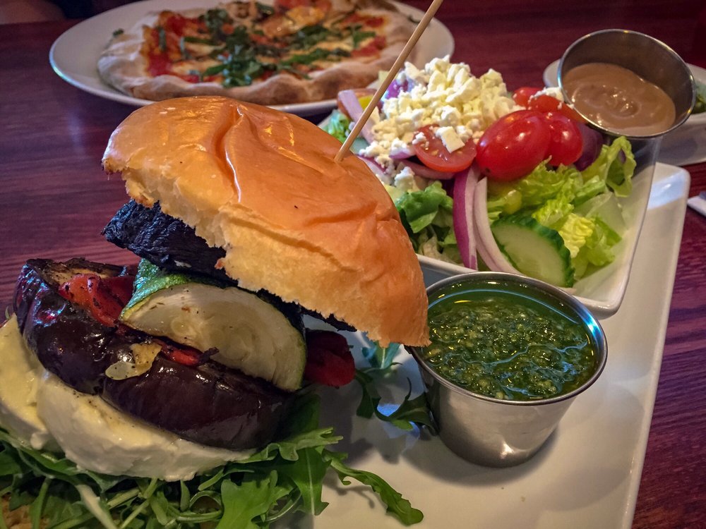 Grilled Vegetable Powerhouse sandwich on a plate, with a salad and a Neapolitan pizza in the background.