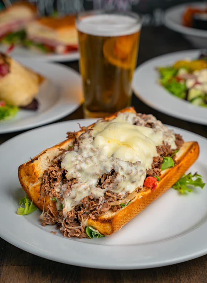 a cheesesteak and a draft beer, with other plates of food in the background