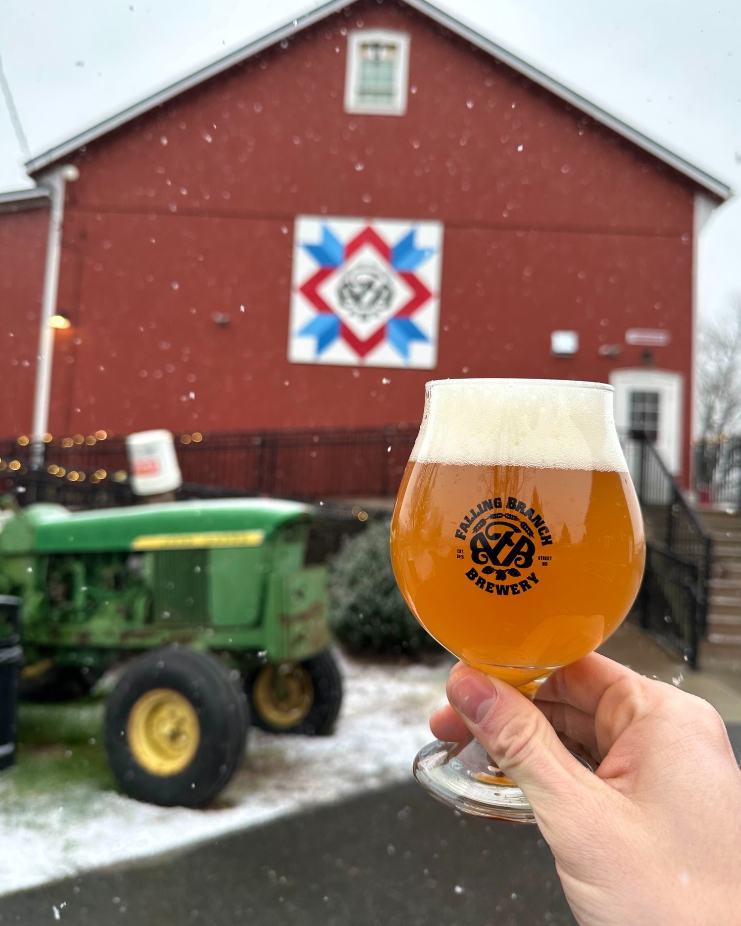 a hand holding a glass of "frostbite pines" beer with a barn quilt and tractor in the background