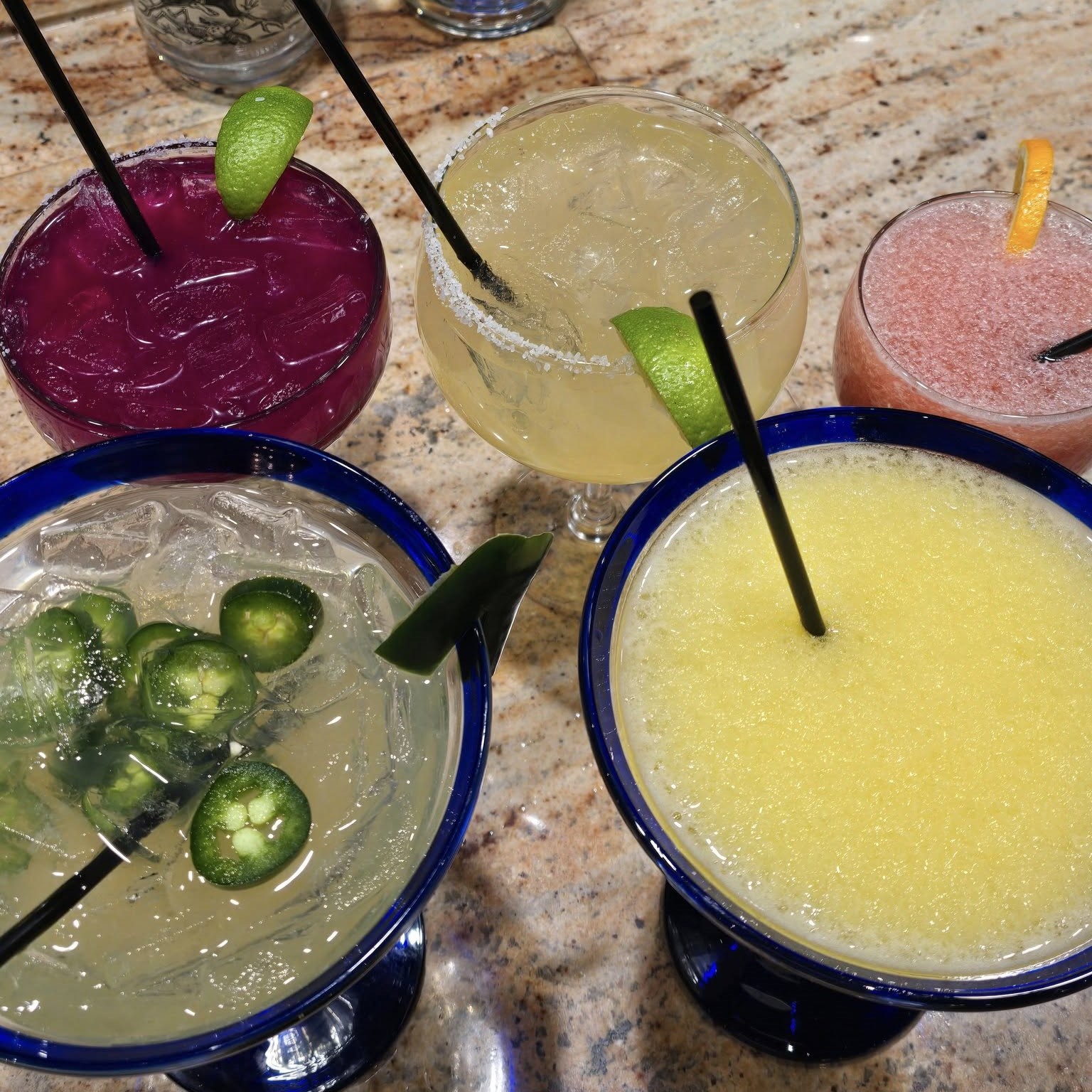 five different colored cocktails, three with lime wedges, one with jalapenos, and one with an orange wheel