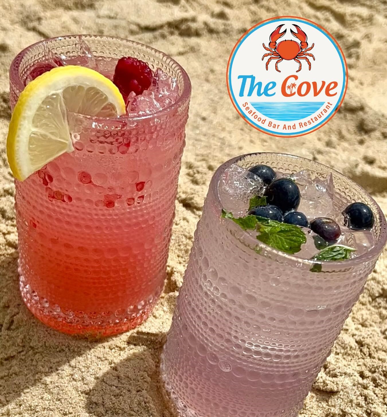 two cocktails sitting on the sand, one red with raspberries and a lemon wedge, the other purple with blueberries and mint