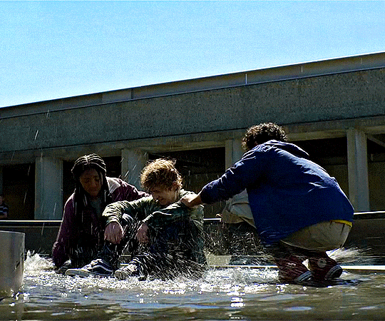 a gif (from the disney+ tv show) of annabeth and grover splashing percy with water in a fountain
